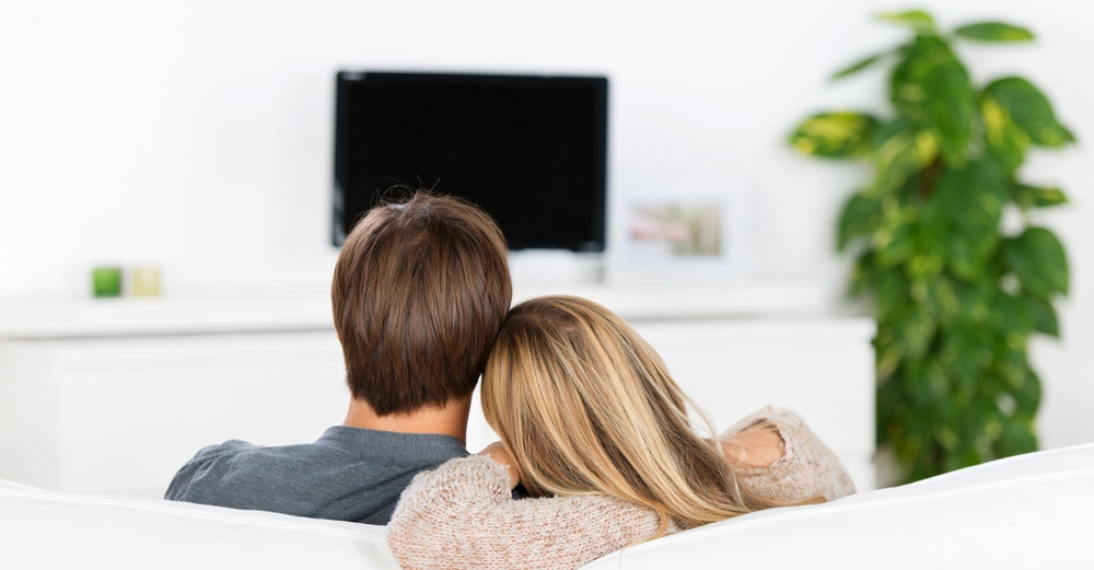 Ein Paar sitzt auf einem weißen Sofa und schaut auf einen ausgeschalteten Fernseher in einem hellen Wohnzimmer mit einer Pflanze und einem Bilderrahmen im Hintergrund.