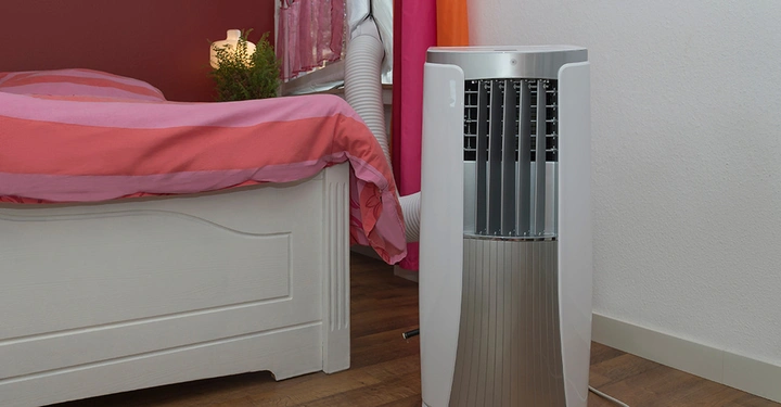 Weißer Luftkühler steht neben einem Bett mit rosa und oranger Bettwäsche in einem Zimmer mit Holzboden und farbenfrohen Vorhängen. Eine Pflanze und ein Licht befinden sich auf dem Nachttisch.