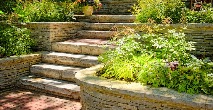 Steintreppe führt durch abgestufte Gartenbeete mit grünen Pflanzen und bunten Blumen umgeben von Steinmauern in einem sonnigen Außenbereich. Ein Blumentopf steht oben auf den Stufen.