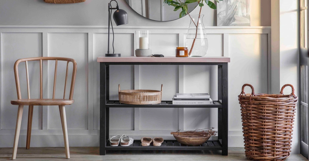 Ein Holzstuhl steht links neben einem schwarzen Metalltisch mit Holzplatte auf dem eine Lampe Kerze und Vase mit Zweig stehen Hinter dem Tisch eine Wand mit Paneelen und rundem Spiegel. Rechts vom Tisch steht ein geflochtener Korb.