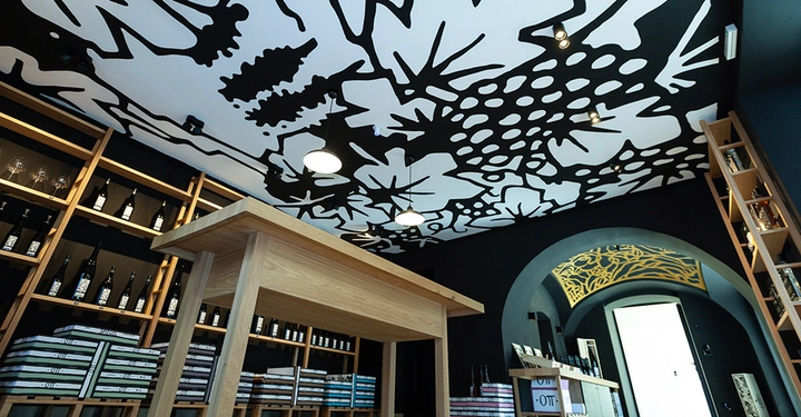 Galerie-Bild 12: Ein hölzerner Tisch steht in einem Raum mit Regalen voller Weinflaschen und Bücher. An der Decke befindet sich ein großflächiges, schwarz-weißes Muster.