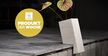 Weiße Vase mit zwei exotischen roten Blumen steht auf einem grauen Tisch in fabrikhallenähnlicher Umgebung. Gelber Kreis mit weißem Kalender und Text: 11 PRODUKT DER WOCHE.