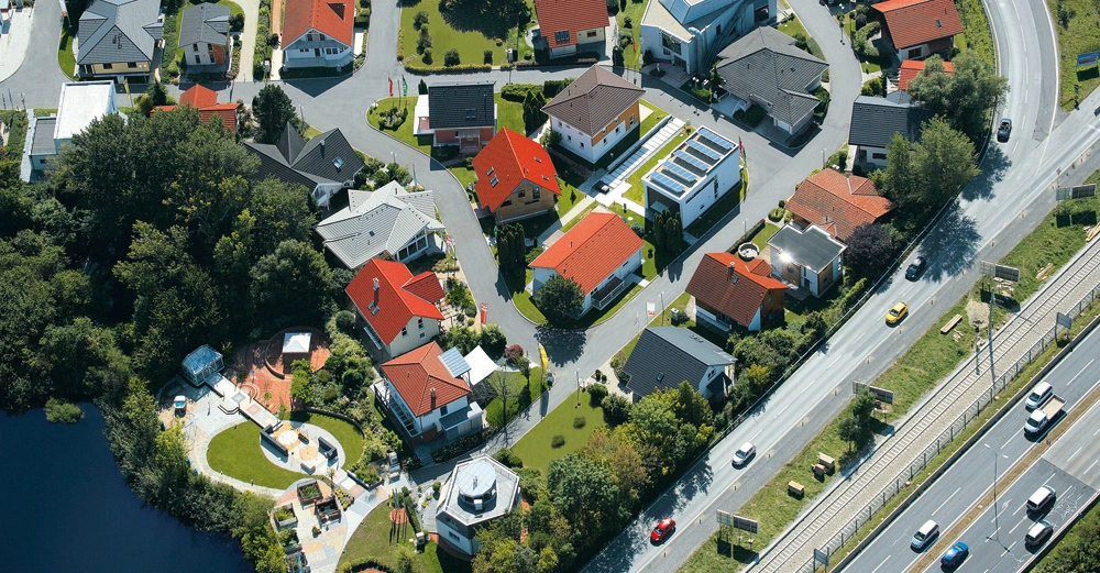 Wohnhäuser stehen in einer kreisförmigen Anordnung in einer modernen Wohnsiedlung, umgeben von Grünflächen und Bäumen links neben einer stark befahrenen Straße mit mehreren Spuren und Autos.