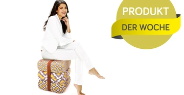 Eine Frau sitzt lächelnd auf stapelbaren Bedruckten Kissen in einem weißen Raum. Rechts im Bild steht in einem gelben Kreis das Text PRODOKT DER WOCHE.