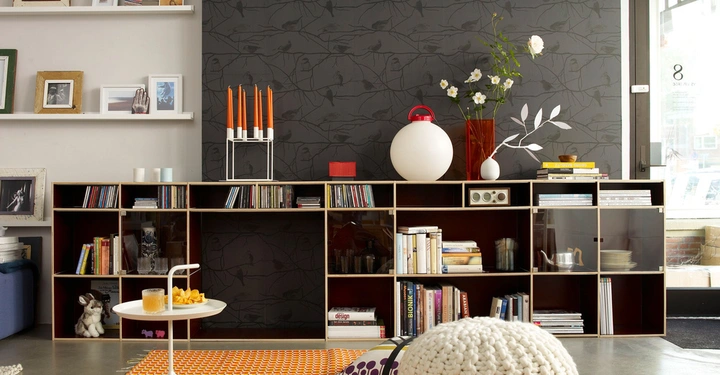 Regal mit Bücher CDs und Dekorationen steht in einem Wohnzimmer vor einer dunkelgrauen Tapete. Ein Couchtisch mit Getränk und Chips steht vorne. Links sind Wandregale mit gerahmten Bildern