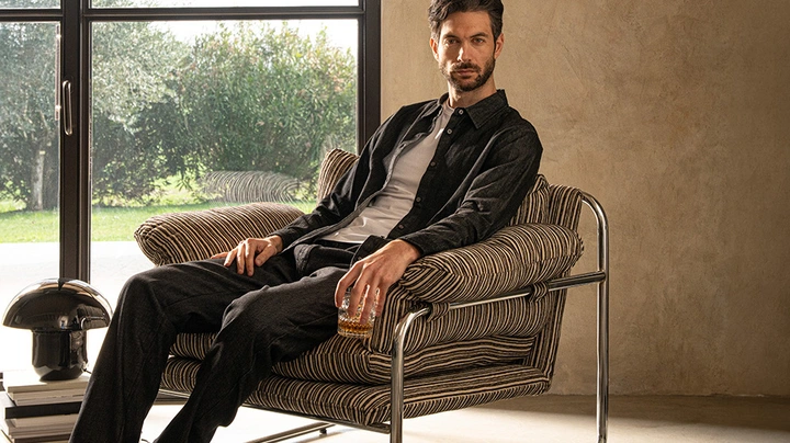 dunkelhaariger Mann mit Bart sitzt mit Whiskeyglas in der Hand in einem Loungesessel und blickt cool in die Kamera