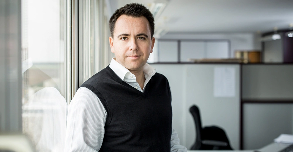 Ein Mann steht an einem Fenster und schaut in die Kamera Büro im Hintergrund mit Schreibtischen und Stühlen Büroflur.