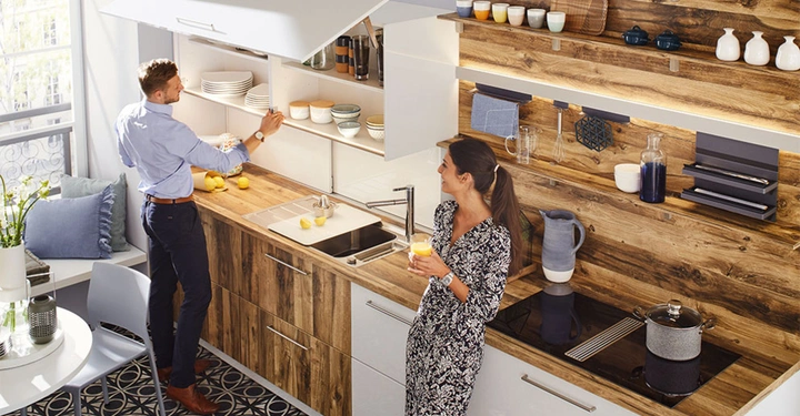 Ein Mann stellt Geschirr in ein Regal eine Frau hält ein Glas Saft in einer modernen Küche mit Holz- und Weißakzenten und verschiedenen Küchenutensilien auf der Arbeitsfläche und an der Wand.