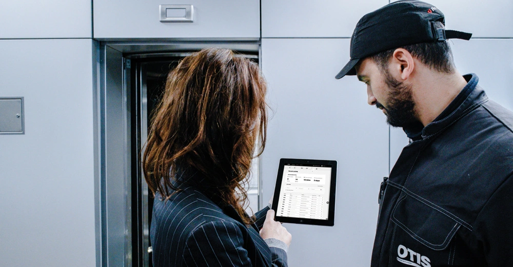 Eine Frau und ein Mann in Arbeitskleidung betrachten ein Tablet in einer modernen Umgebung mit einem Aufzug im Hintergrund. Der Text auf dem Tablet ist schwer lesbar.