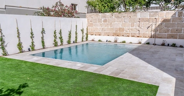 Ein rechteckiger Swimmingpool ruht still in einem minimalistisch gestalteten Garten umgeben von einem Ziegelsteinmauer und grünen Pflanzen während der Boden aus sauberem, hellem Stein und gepflegtem Kunstrasen besteht.