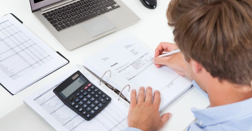 Eine Person schreibt an einem Schreibtisch mit einem Laptop und einer Maus Rechnungen in einen Ordner und nutzt dabei einen Taschenrechner.