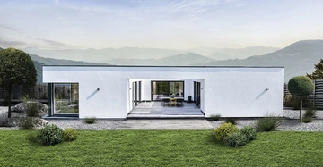 Modernes weißes Einfamilienhaus mit Flachdach steht ruhig inmitten eines gepflegten Gartens vor einer bergigen Landschaft. Im Vordergrund befindet sich eine Terrasse mit Tisch und Stühlen.