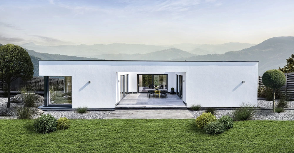 Modernes weißes Einfamilienhaus mit Flachdach steht ruhig inmitten eines gepflegten Gartens vor einer bergigen Landschaft. Im Vordergrund befindet sich eine Terrasse mit Tisch und Stühlen.