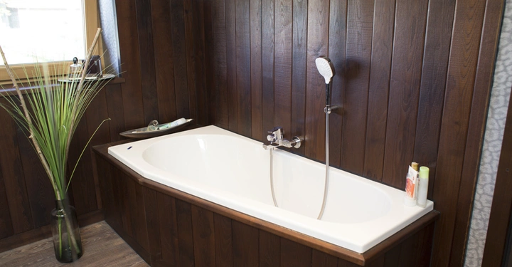 Galerie-Bild 4: Weiße Badewanne mit Handbrause an der Wand montiert holzverkleidete Wände eine Vase mit hohen grünen Pflanzen am Fenster ein Regal mit Badeutensilien Fliesenboden helle Wände darüber