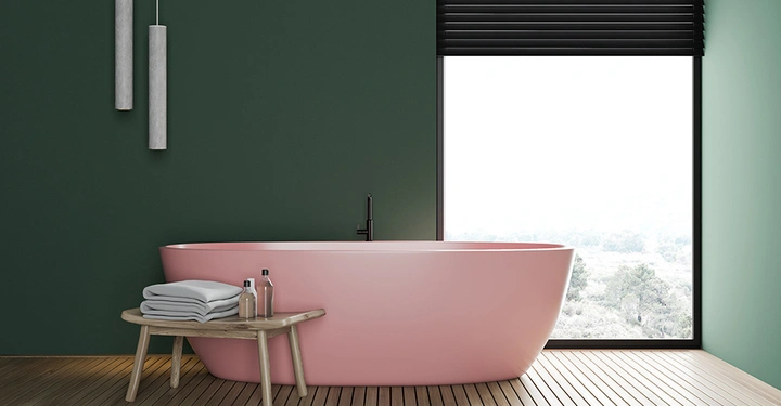 Rosa Badewanne steht in modernem Badezimmer neben kleinem Holztisch mit Handtüchern und Flaschen Vor großem Fenster mit schwarzer Jalousie Hängelampen an dunkelgrüner Wand Holzboden fügt Wärme hinzu
