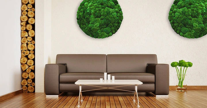 Braunes Sofa steht vor einer Wand mit zwei grünen Moosdekorationen linkerhand gestapeltes Brennholz Holzboden weißer minimalistischer Couchtisch mit Dekoration grüne Pflanzen daneben in Vase