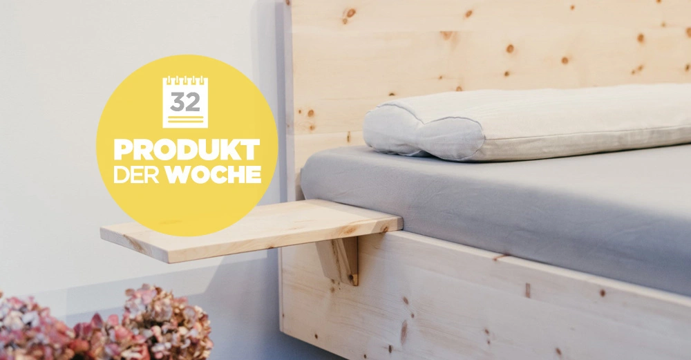 Ein hölzernes Bett mit grauer Bettwäsche und einem weißen Kissen steht in einem hellen Raum. Ein gelbes Symbol zeigt Text '32 PRODUKT DER WOCHE'. Links unten sind getrocknete Blumen.