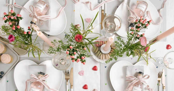 Gedeckter Tisch mit einer Flasche Mirabeau auf einem Herz-Teller, rosa Blumen in Gläsern und brennenden Kerzen in der Mitte, umgeben von Macarons und Cupcakes auf hellem Tischtuch.