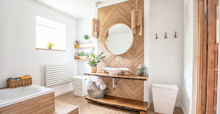 Rundes Spiegel über Waschtisch mit weißen Becken in modernem Badezimmer hölzerne Wand und Böden weiße Wände links ein Fenster rechts Haken und Korb Pflanzen Dekorationen und Regale mit Gegenständen.