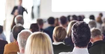 Menschen sitzen zugewandt einem Präsentator der vorne steht und spricht in einem Konferenzraum erhöht durch unscharfe Darstellung des Präsentators im Hintergrund.