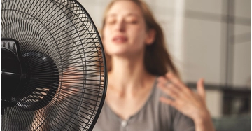Schwarzer Standventilator im Vordergrund kühlt eine Person in einem hellen Wohnraum während eines heißen Sommertages.