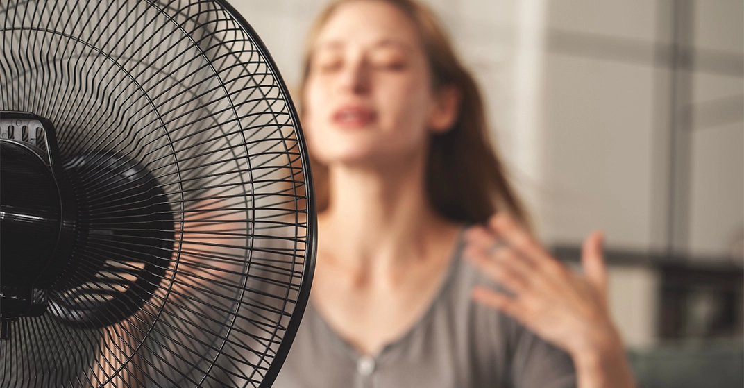 Schwarzer Standventilator im Vordergrund kühlt eine Person in einem hellen Wohnraum während eines heißen Sommertages.