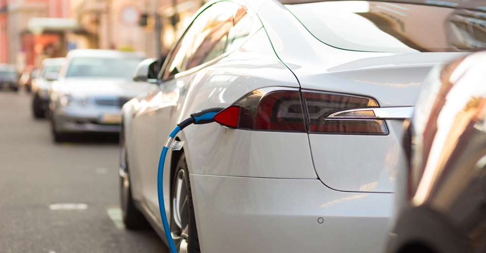 Weißes Elektroauto lädt mit blauem Ladekabel am Straßenrand Mehrere Autos parken im Hintergrund in einer städtischen Umgebung an der Straße