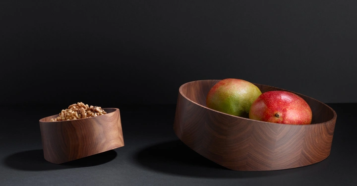 Zwei Holfschalen enthalten Walnüsse und Mangos, die auf einem flachen, schwarzen Hintergrund liegen. Die linke Schale ist kleiner und enthält Walnüsse, die rechte größere Schale enthält zwei Mangos.