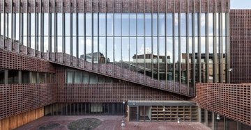 Ein modernes Gebäude mit großen Glasfenstern und roten Backsteinwänden steht in einem Innenhof. Das Design enthält vertikale Metallstreben und diagonale Strukturen im oberen Bereich.