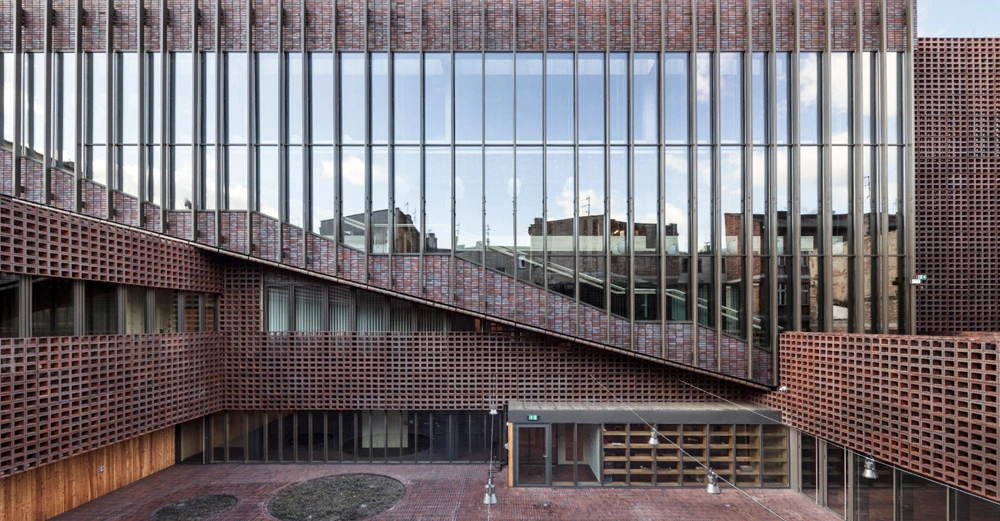 Ein modernes Gebäude mit großen Glasfenstern und roten Backsteinwänden steht in einem Innenhof. Das Design enthält vertikale Metallstreben und diagonale Strukturen im oberen Bereich.