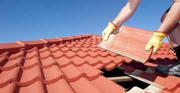 Person setzt Ziegel auf ein Dach in einer sonnigen Umgebung mit klarem blauem Himmel.