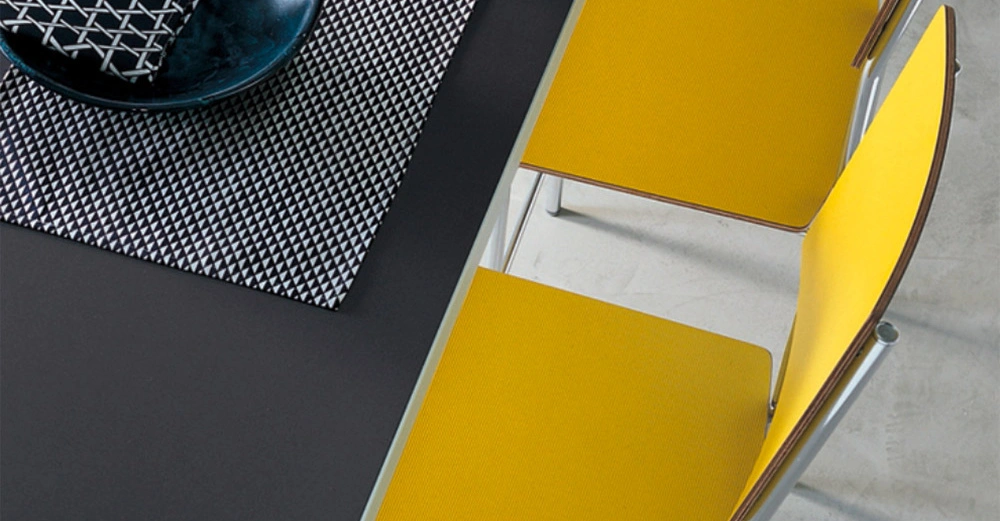 Gelbe Stühle stehen an einem dunkelgrauen Tisch mit geometrisch gemustertem Platzdeckchen und einer dunkelblauen Schale darauf. Der Raum hat einen hellen Boden.