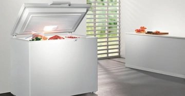 Gefrierschrank mit offenem Deckel enthält gefrorene Lebensmittel in Tüten steht in einem hellen Raum mit Lamellenfenster und einer weißen Anrichte auf der Obst und ein Schneidebrett liegen
