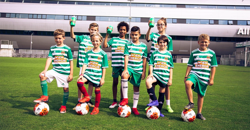Kinder posieren auf einem Fußballfeld. Sie tragen grün-weiß gestreifte Trikots mit der Aufschrift WIEN ENERGIE und heben Wasserflaschen. Im Hintergrund ist ein modernes Gebäude zu sehen.