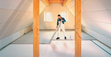 Handwerker verlegt Bodenplatten in einem hellen Dachboden mit schrägen Wänden und zwei kleinen Fenstern auf der Stirnseite Holzträger und Balken umgeben den Raum.