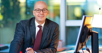 Ein Mann im Anzug sitzt an einem Schreibtisch und schaut in die Kamera im Hintergrund befinden sich ein Monitor eine Tischlampe und ein großes Fenster.