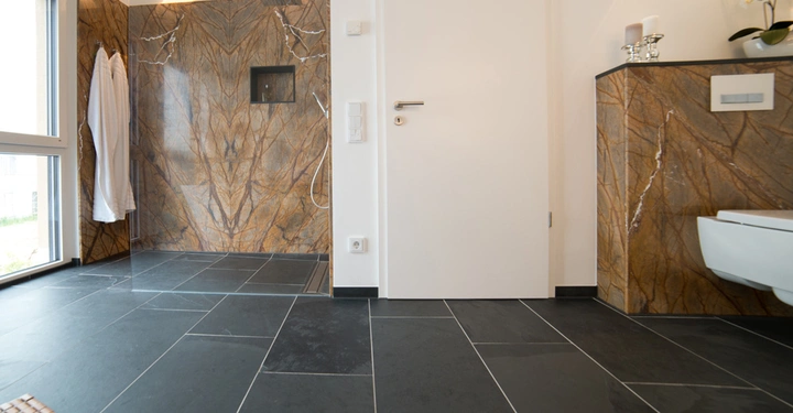 Duschbereich mit Marmorwänden und Regenduschkopf links neben einer weißen Tür. Umgebung: Badezimmer mit dunklen Fliesen, WC und Waschbecken rechts, Fenster links, zwei aufgehängte Bademäntel.