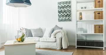 Weißes Sofa mit gemusterten Kissen steht vor weißer Wand und Regal voll mit Körben und Büchern darunter ist ein kleiner Tisch mit einem Obstkorb dekoriert der Raum ist hell erleuchtet