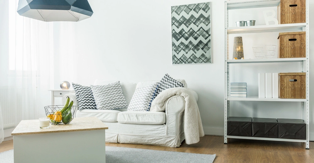 Weißes Sofa mit gemusterten Kissen steht vor weißer Wand und Regal voll mit Körben und Büchern darunter ist ein kleiner Tisch mit einem Obstkorb dekoriert der Raum ist hell erleuchtet