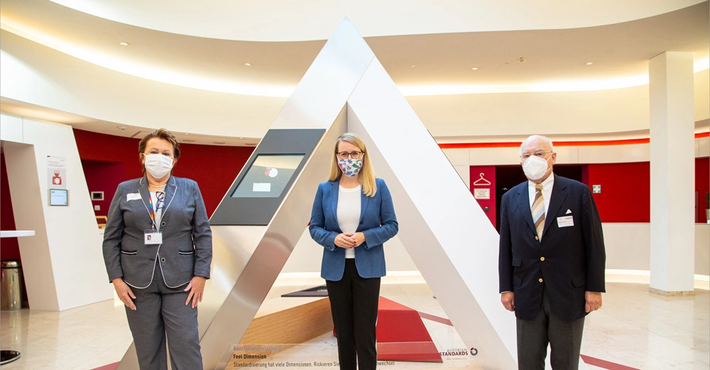 Drei Personen stehen mit Masken vor einer großen dreieckigen Installation in einer hellen, modernen Lobby mit roten Akzenten. Text auf der Installation Feell Dimension. Standardisierung hat viele Dimensionen Risikieren Sie mehr und entdecken Sie Standards mit Unterschieden heute und morgen.