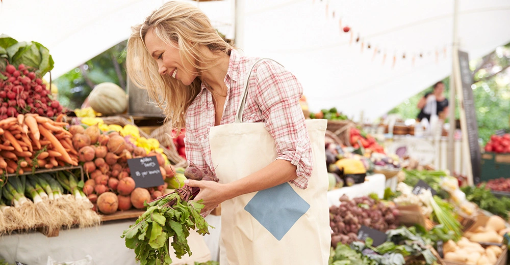 Eine Frau trägt eine Einkaufstasche und wählt frisches Gemüse auf einem lebhaften Bauernmarkt aus. Im Hintergrund sind mehrere Stände mit bunten Produkten und anderen Kunden zu sehen.