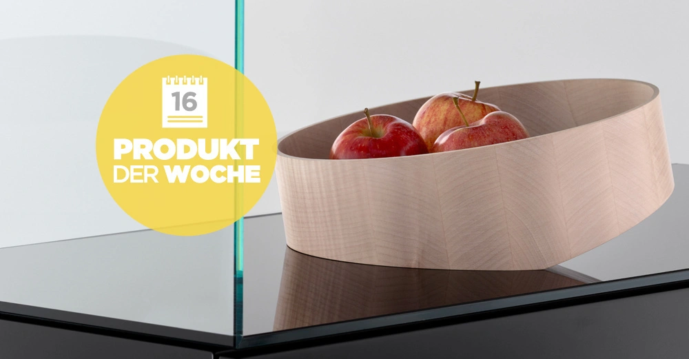 Holzschale mit roten Äpfeln steht auf einer schwarzen glänzenden Oberfläche mit einer Glasplatte. Ein gelber Kreis mit Text "16 PRODUKT DER WOCHE" ist links sichtbar.