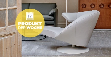 Ein moderner weißer Stuhl steht auf einem grauen Teppich vor einem grünen Sofa und braunen Holzschränken. Text: 17 PRODUKT DER WOCHE.