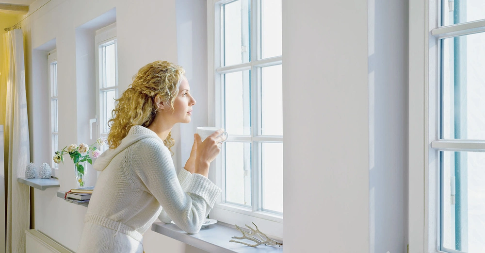 Frau hält eine Tasse und schaut aus einem Fenster in einem hellen Zimmer mit weißen Wänden und dekorierten Fensterbänken.