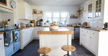 Eine weiße Salatschüssel mit Orangen steht auf einer Kücheninsel; Hocker davor; umgeben von weißen Schränken und diversen Küchengeräten in einer hellen Küche mit großen Fenstern. Text: "Stuff".