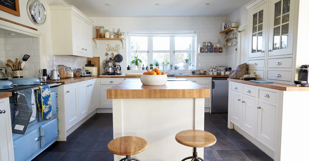 Eine weiße Salatschüssel mit Orangen steht auf einer Kücheninsel; Hocker davor; umgeben von weißen Schränken und diversen Küchengeräten in einer hellen Küche mit großen Fenstern. Text: "Stuff".