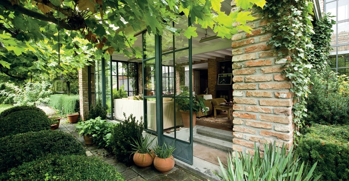 Eine verglaste Terrassentür steht offen und führt in einen gemütlich eingerichteten Wohnbereich mit Pflanzen auf einem Steinfundament. Draußen umgeben grüne Blätter und üppige Vegetation das Haus.