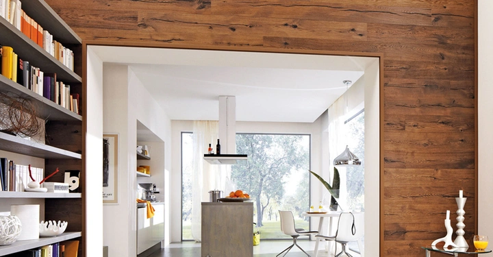 Raum mit dunklen Holzwänden enthält Regale voller Bücher. Mittig befindet sich eine Durchgangstür zur hellen, modernen Küche, in der eine Kochinsel und ein Esstisch mit Stühlen stehen.
