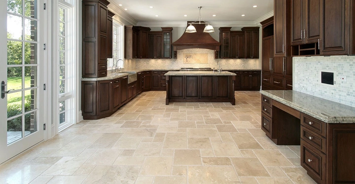 Große luxuriöse Küche mit dunklen Holzmöbeln und hellen Fliesenböden leere Kochinsel in der Mitte Einbaugeräte und helles Tageslicht durch große Fensterflügeltüren Öffnung zum grünem Garten