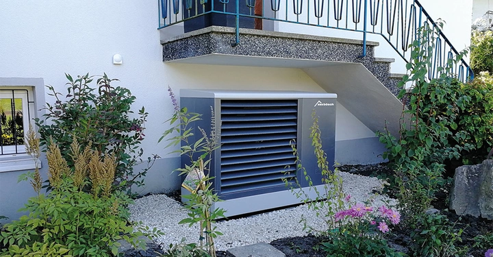 Ein graues Lüftungsgerät steht ruhig im Garten neben einem weißen Haus unter einer Treppe und ist von verschiedenen Pflanzen umgeben. Text: Stiebel Eltron.